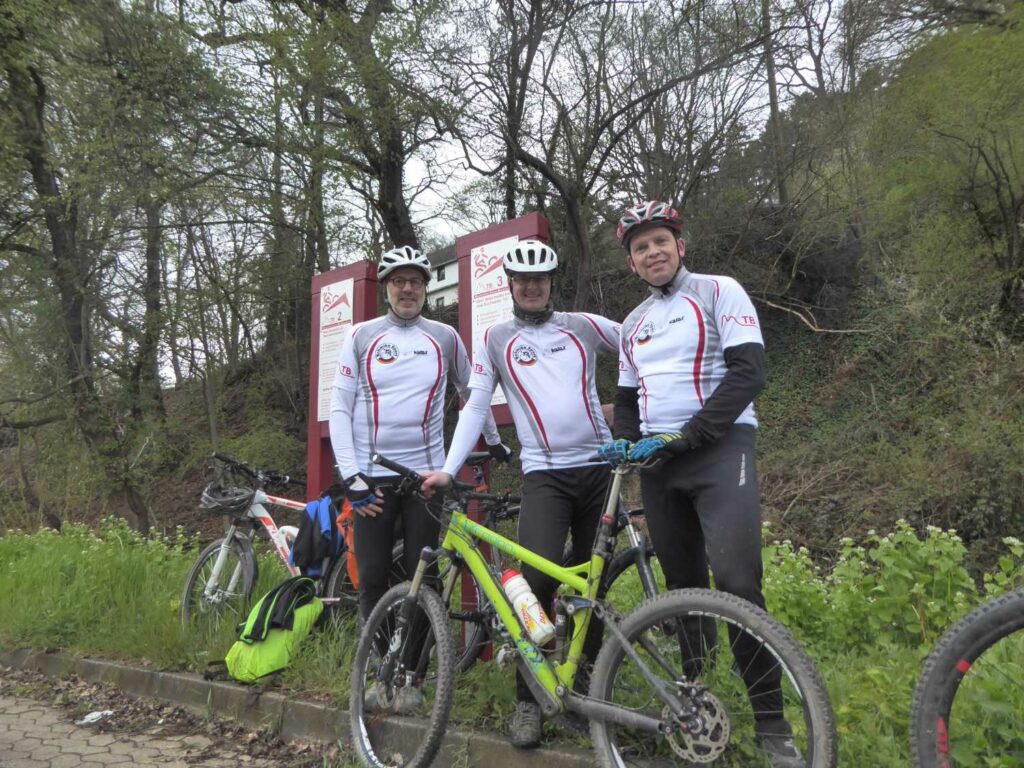 Drei Männer mit Helm und Fahrradkleidung stehen mit ihren Bikes im Wald vor einer Infotafel zu Mountaibikestrecken